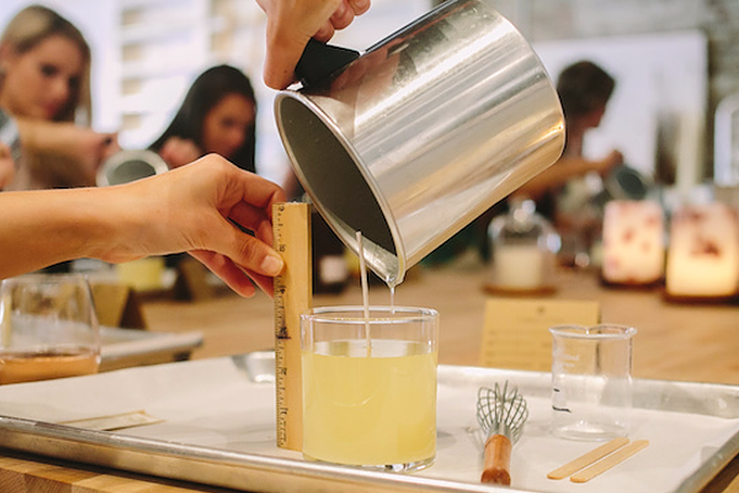 Person pouring a drink from a metal pitcher into a glass on a wooden table with other people in the background.